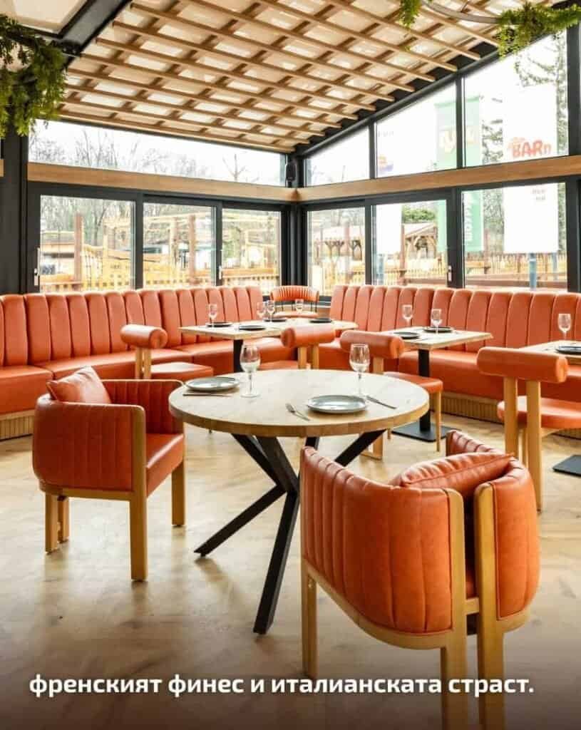 Стилна вътрешна част на ресторант с уютни оранжеви столове и големи прозорци.