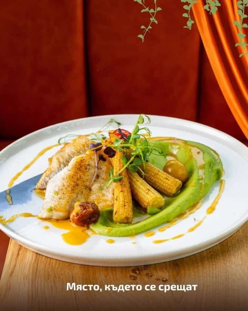 Прясна риба с гарнитура от зеленчуци и сос, сервирана на бяла чиния в уютна обстановка.